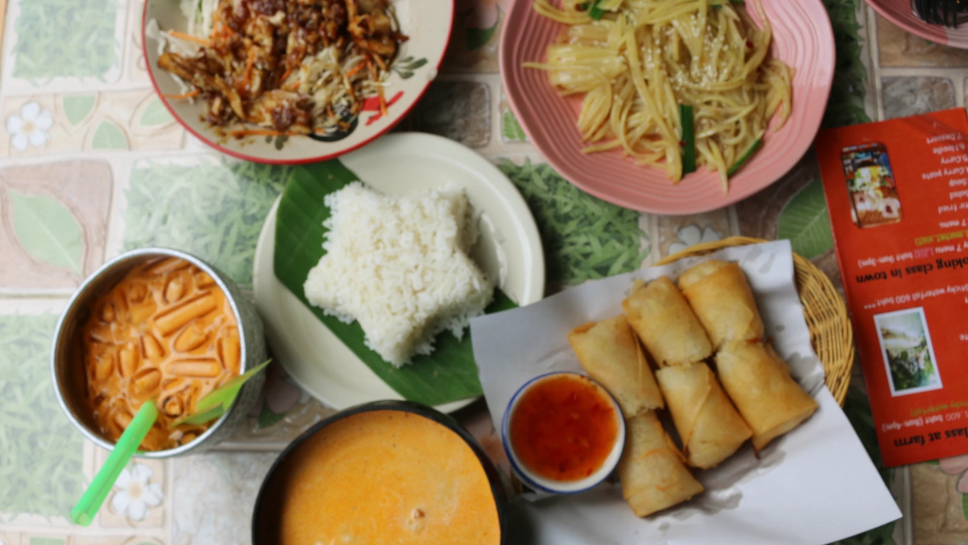 Table of food featuring Thai milk tea, spring rolls, boiled rice in the shape of a star, Thai curry, stir fried potatoes and chicken